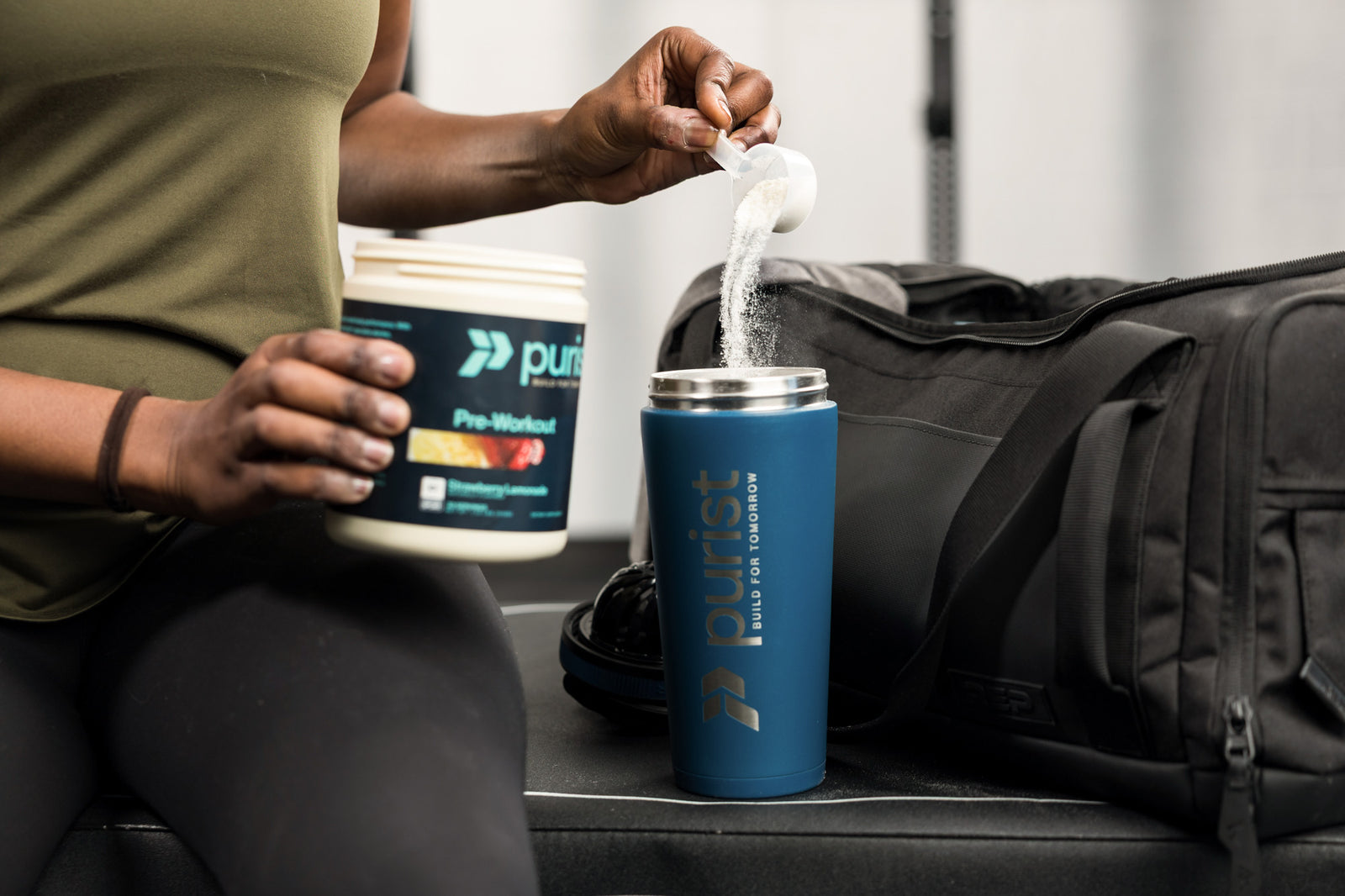 Person pouring powder from a Purist container into a blue shaker bottle with a gym bag in the background.